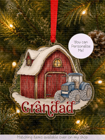 A personalized Christmas ornament shaped like a red barn with a blue tractor and snow, hanging on a Christmas tree. The ornament has the name 'Gréndad' written in red script and a white circular sticker that says 'You can Personalise Me!'.