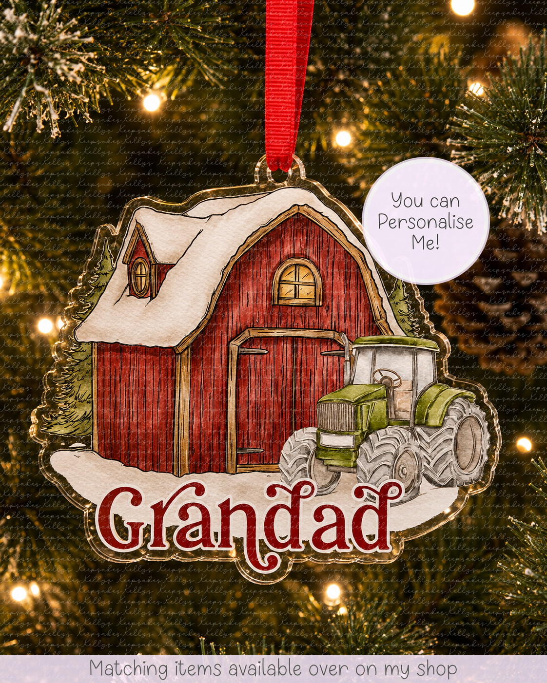 A personalized Christmas ornament shaped like a red barn with a green tractor and snow, hanging on a Christmas tree with warm lights. The ornament has the word 'Grandad' in red script and a white circle that says 'You can Personalise Me!'.