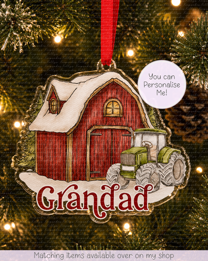 A personalized Christmas ornament shaped like a red barn with a green tractor and snow, hanging on a Christmas tree with warm lights. The ornament has the word 'Grandad' in red script and a white circle that says 'You can Personalise Me!'.