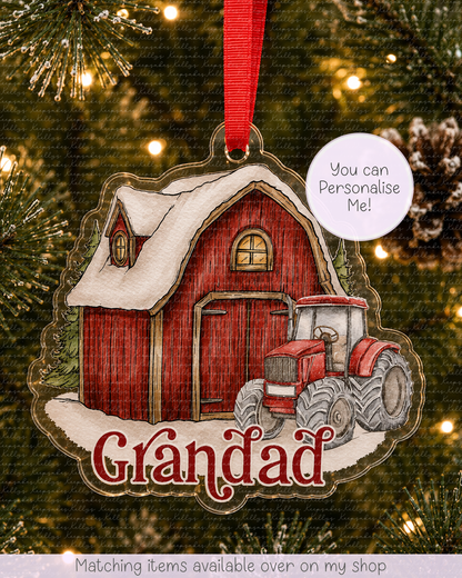 A personalized Christmas ornament shaped like a red barn with a tractor and snow, hanging on a Christmas tree. The ornament has the name 'Grandad' written on it and a red ribbon at the top.